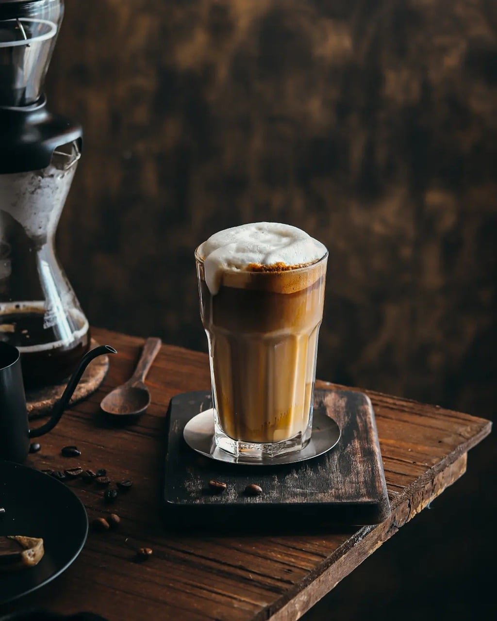 iced latte macchiato, layered espresso, milk foam, wooden spoon, chemex, coffee beans, dark textured table, metal saucer, wooden tabletop, rustic coffee maker, artistic drink, slow living, cozy morning, coffee shadow effect, warm light, wood texture, shadow play, afternoon coffee time, aesthetic photography, barista creation